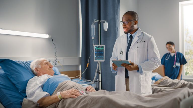 Young male doctor speaks with older male patient at his bedside in a hospital