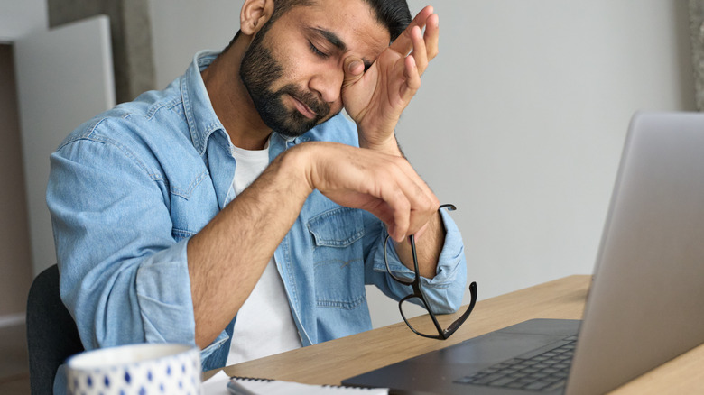 Man sitting with laptop, holding glasses and rubbing eyes