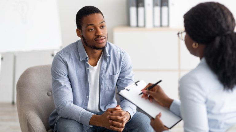 Man sits before a therapist holding a clipboard in a therapy session